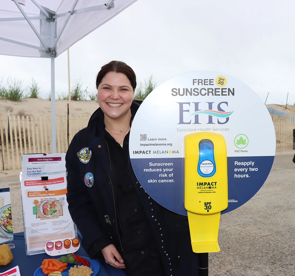 Shoni Rozenberg Seide, DO, FAAD, Associate Dermatology Program Director at Episcopal Health Services stands by one of the sunscreen dispensers EHS is providing to Rockaway beachgoers in partnership with IMPACT Melanoma.